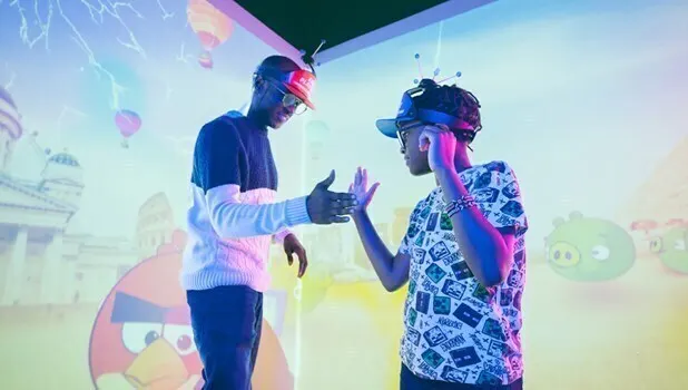 2 black boys wearing VR headsets inside a room with images projected on the walls. 