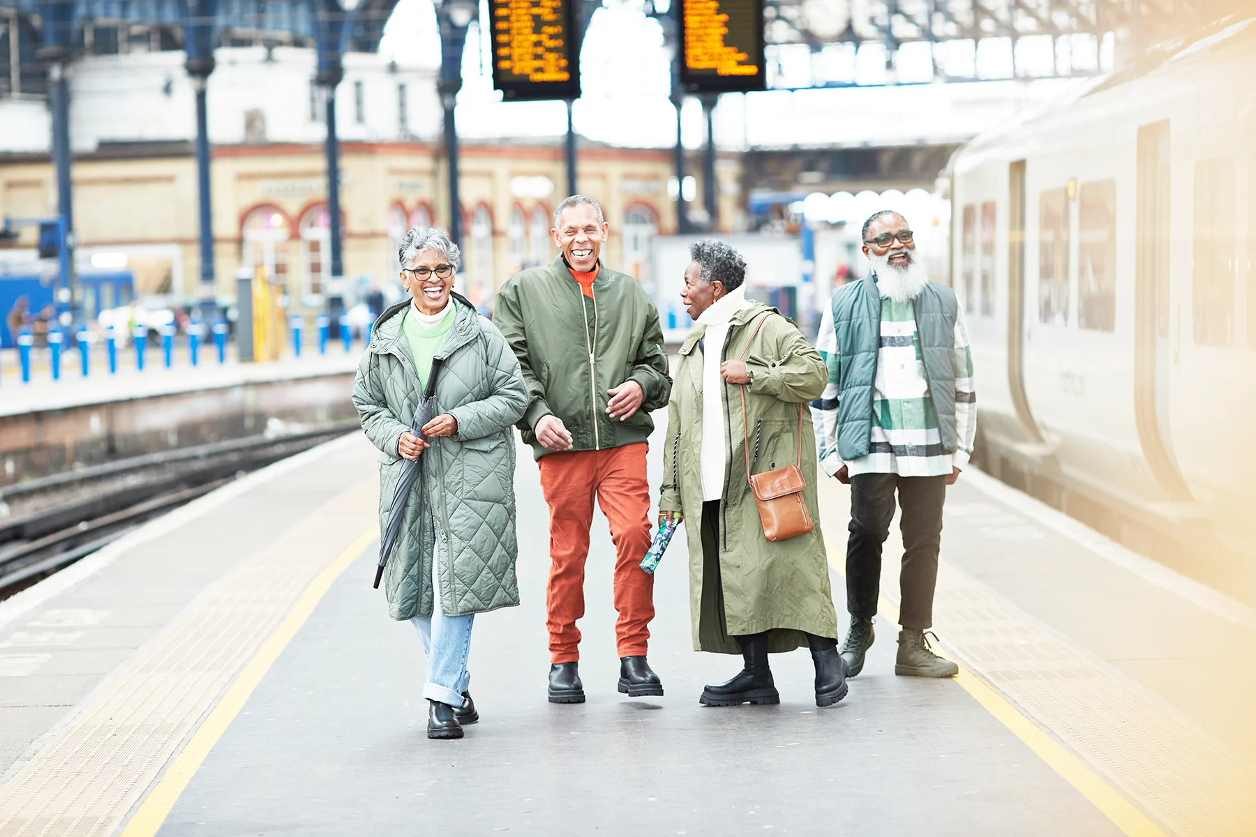 2 senior Black men and 2 senior Black women standing laughing on a station platform with a train on the tracks next to them.