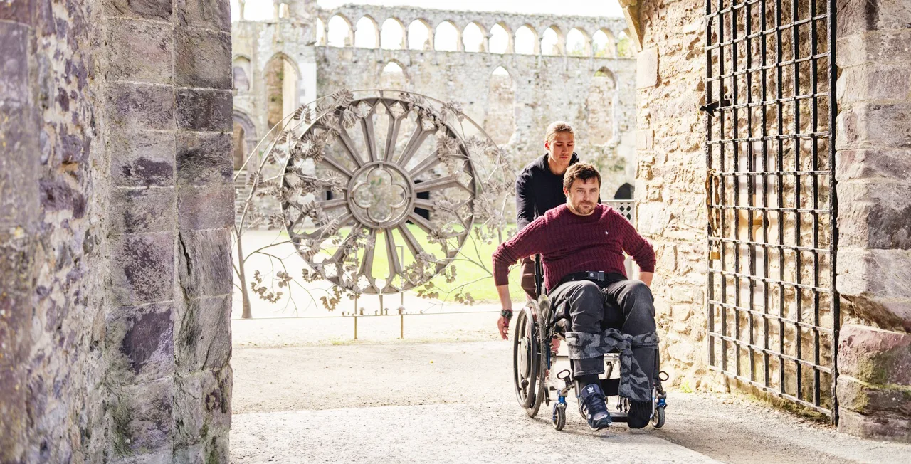 A white man walking behind a white man in a wheelchair, in the ruins of an ancient building. 