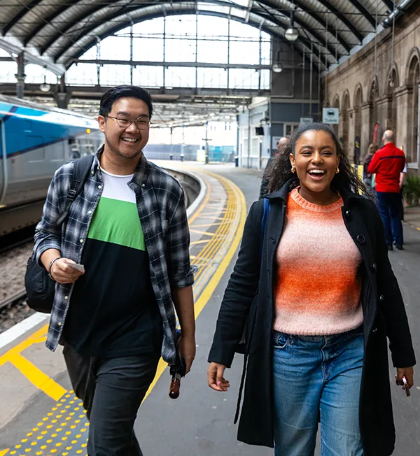 2 young people, a South Asian man and a Black woman, dressed in casual clothes and laughing and smiling as they walk along a platform at a train station. 