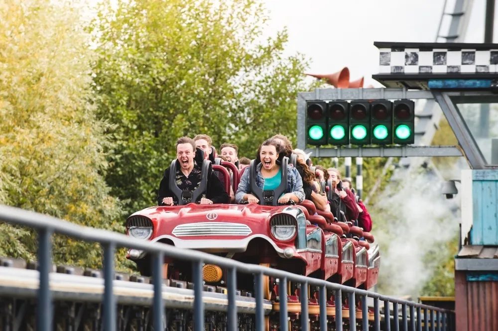 People visibly screaming with excitement while locked into a rollercoaster carriage under a 'go' sign, with trees to the left.