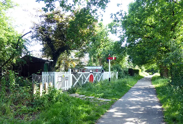A rural footpath with a white railway crossing gate, surrounded by lush green trees and vegetation on a sunny day.