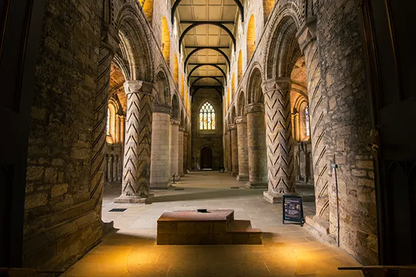 Inside an ancient stone abbey, with columns supporting arches around a central walkway, with stained glass windows on all sides. 