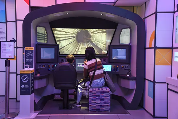 A woman and a boy seen from behind, sitting in a simulator of a London Underground driver's cab. 
