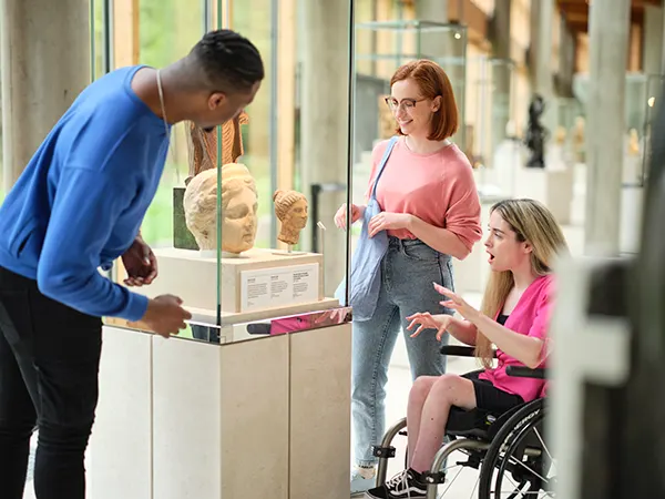 A Black man and 2 white woman, one of whom is a wheelchair user, looking at a bust of a woman's head in a glass museum display cabinet.