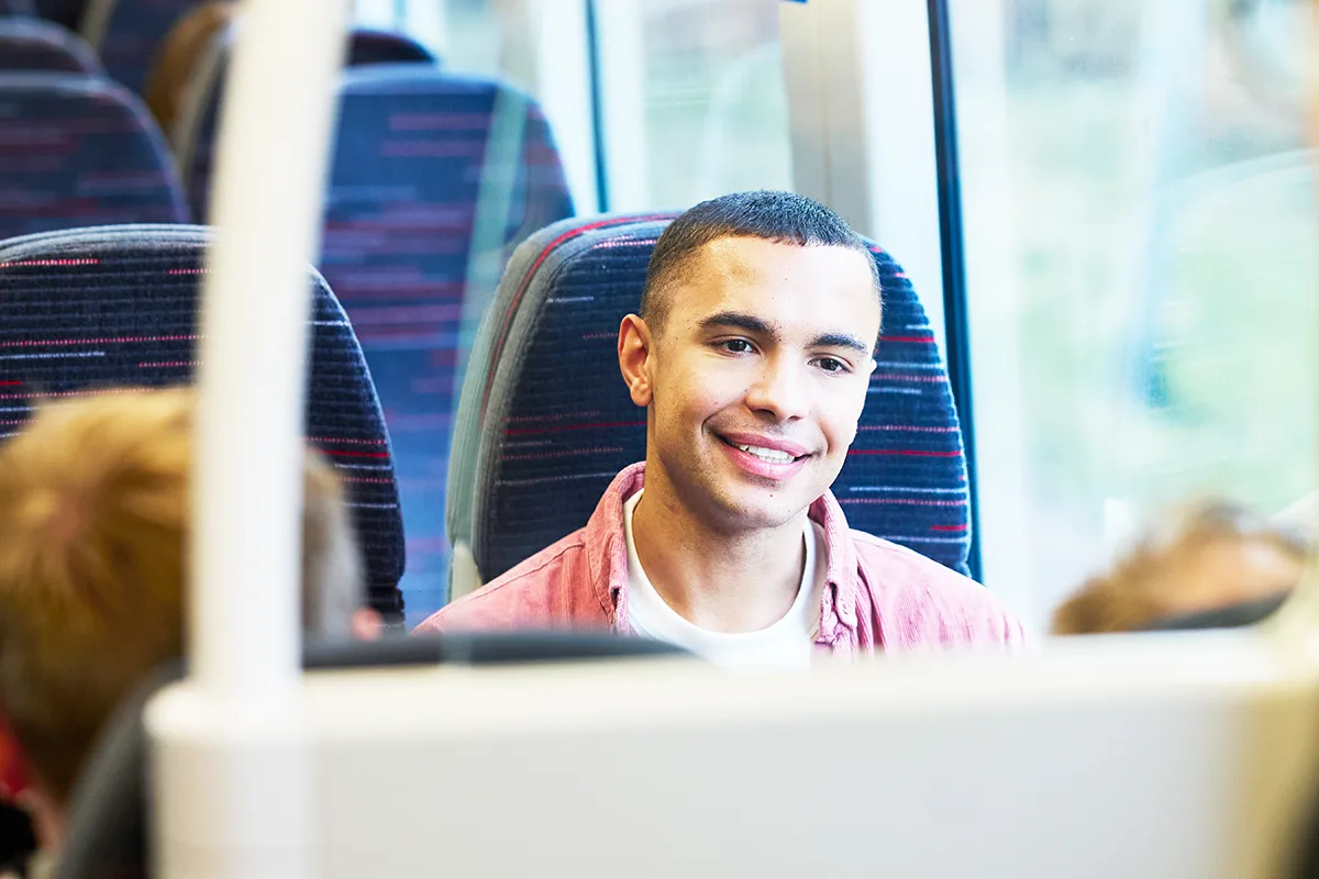 A young man in a pink shirt smiling while seated at a table on a train with other passengers.
