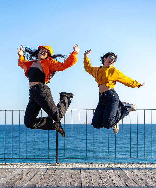 Two people in bright orange and yellow jackets jumping joyfully on a seaside pier against a clear blue sky.