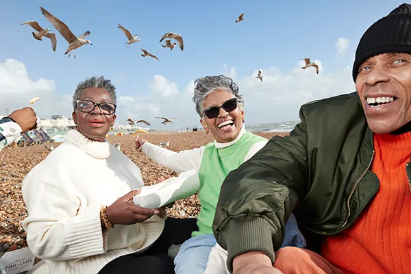 Three Black seniors enjoying a day at the beach with seagulls flying overhead against a bright blue sky.