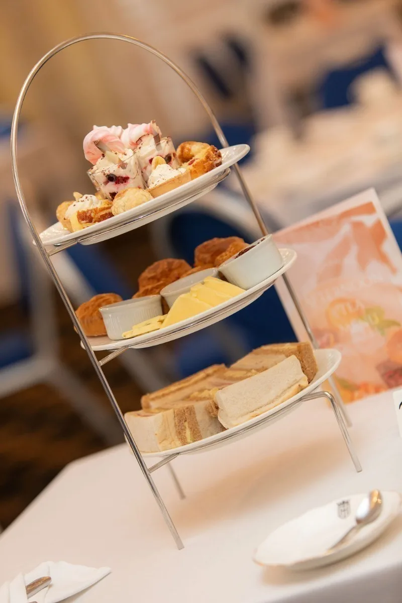 A cake stand set for afternoon tea with finger sandwiches, scones and cakes on top of a white tablecloth. 