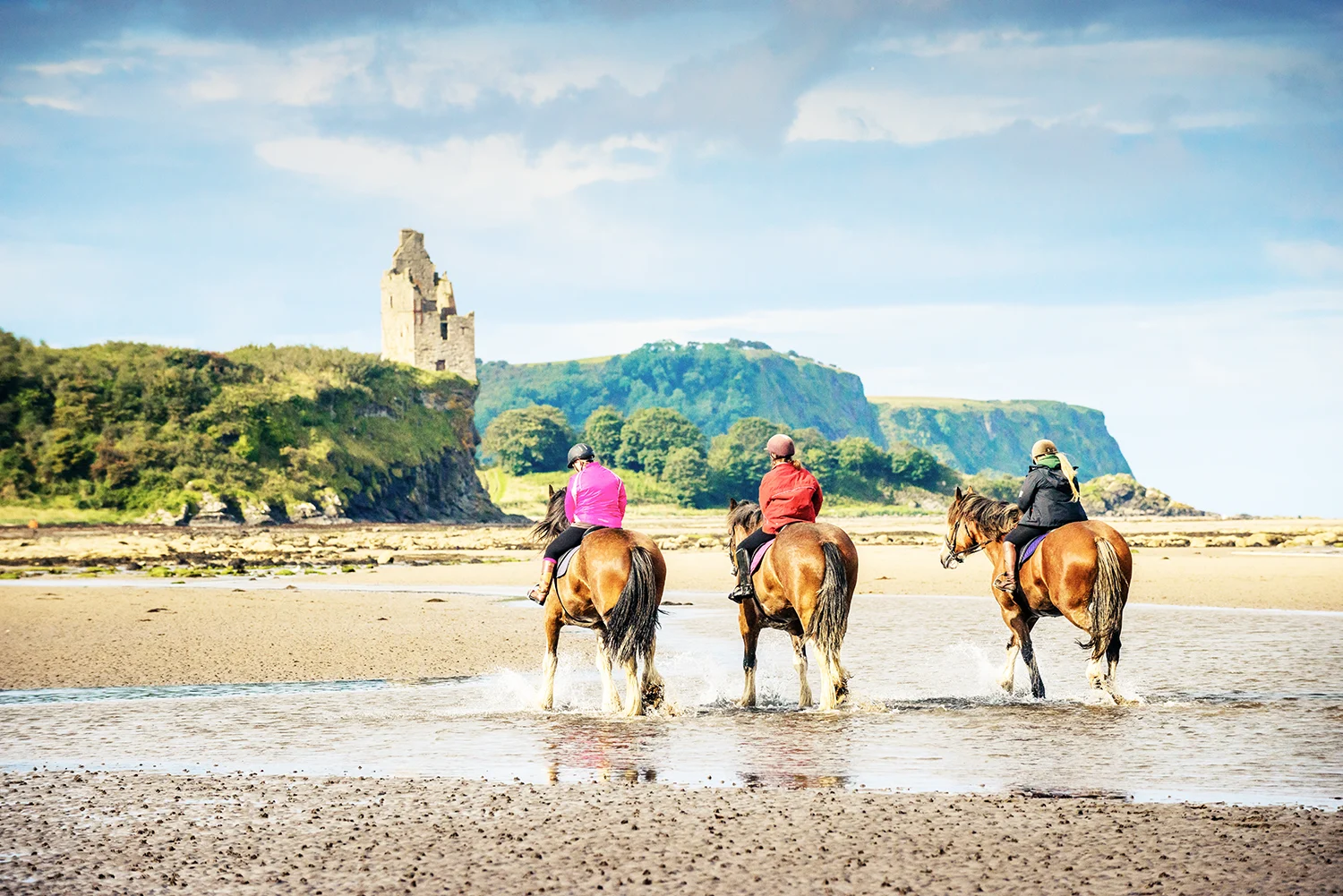 3 horse riders viewed from behind on a sandy beach with large pools, green hillside with the ruins of an old building is visible in the distance. 