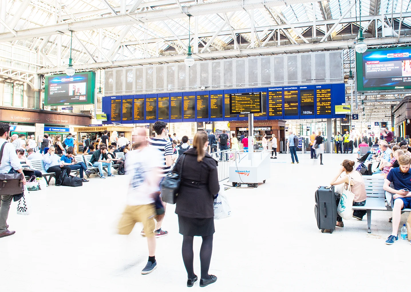 Passengers standing and sitting on a busy station concourse with departure boards visible in the background. 