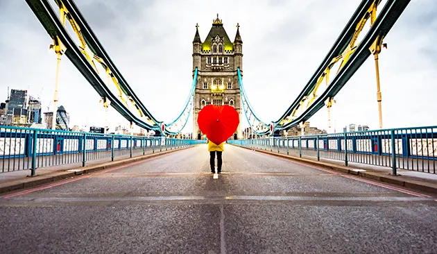 A person standing behind a red heart-shaped umbrella in the middle of Tower Bridge looking towards one of the towers.