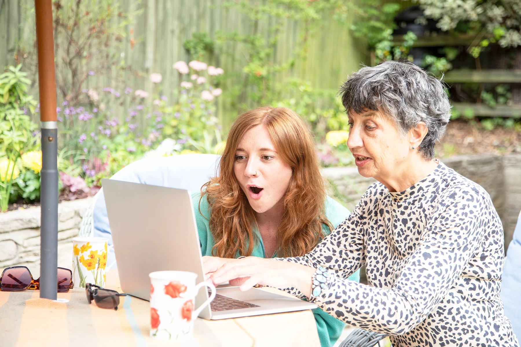 A senior white woman with short grey hair sitting at a garden table with a young woman with long red hair, they are looking at a laptop and the younger woman is gasping with surprise