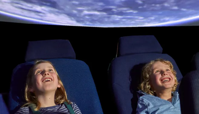 2 white children with blonde hair look up in awe at the ceiling of a planetarium.