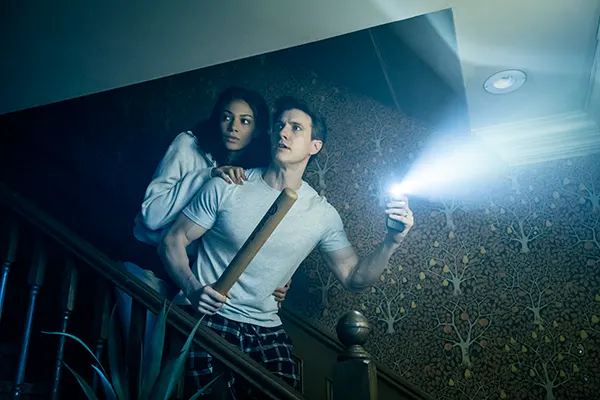 Blue-tinted horror scene with a Black woman and a white man looking frightened on a dark staircase. He is holding his phone and using the torch, and has a baseball bat in the other hand, while she shelters behind him. 
