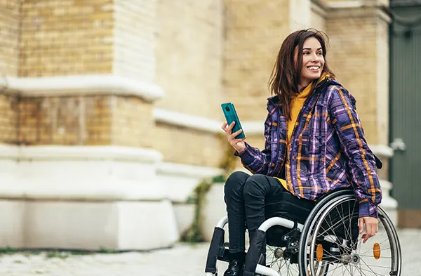A smiling white woman with dark hair, wearing a brightly patterned purple jacket, in a wheelchair and holding a mobile phone.