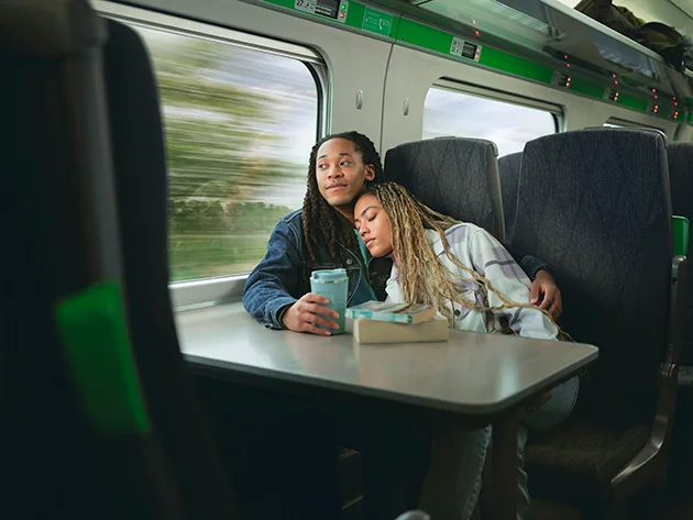 A Black man and Black woman sitting next to each other on a train, he is looking out the window and holding a travel mug while she leans on his shoulder with her eyes closed. 