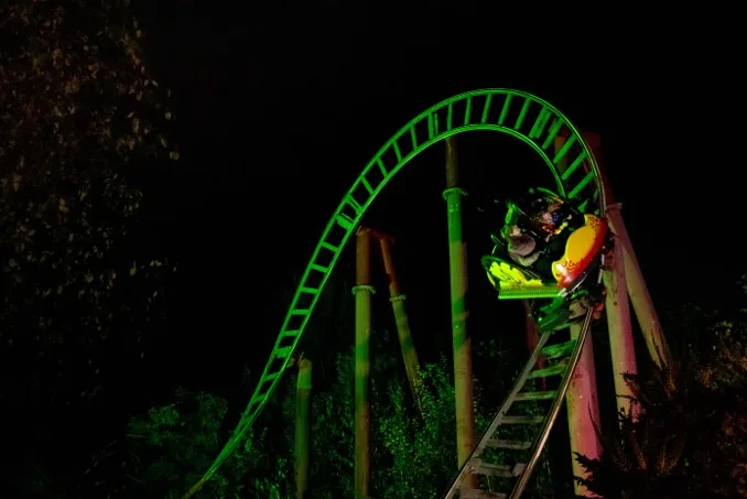 A green-lit roller coaster loop at night, with riders in a colourful cart ascending the illuminated track against a dark sky.