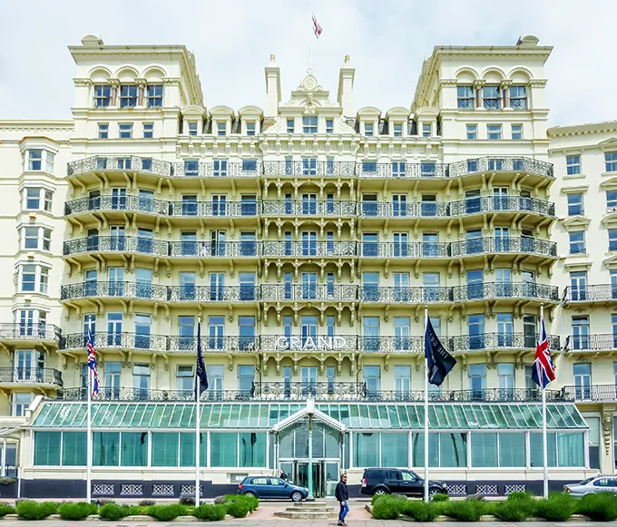 The front of the Grand Hotel in Brighton, a white building of several storeys, with a glass conservatory across the ground floor and entrance. 