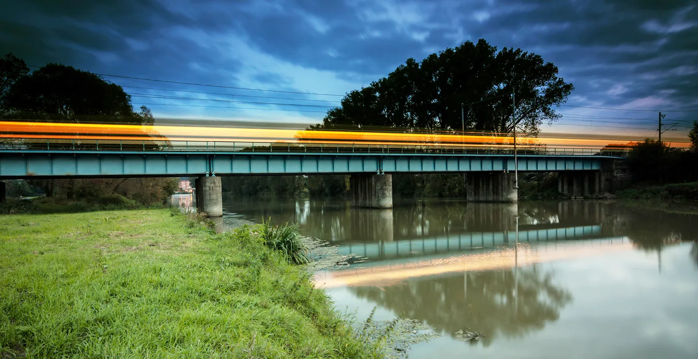 A blurred image of an illuminated, fast-moving train going over a bridge at dusk.