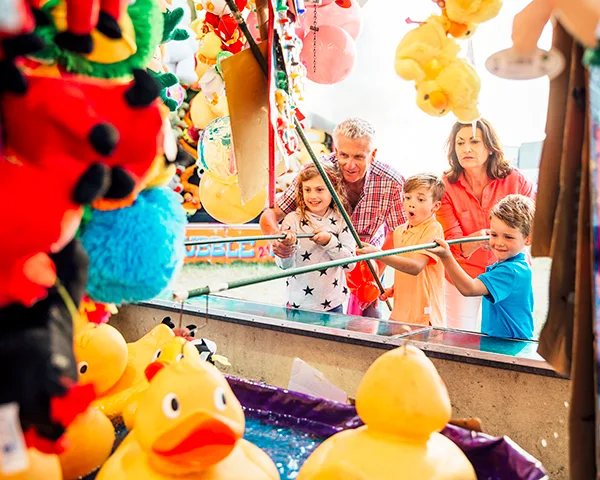 Family enjoying a carnival game with yellow rubber ducks, surrounded by colourful stuffed toy prizes.