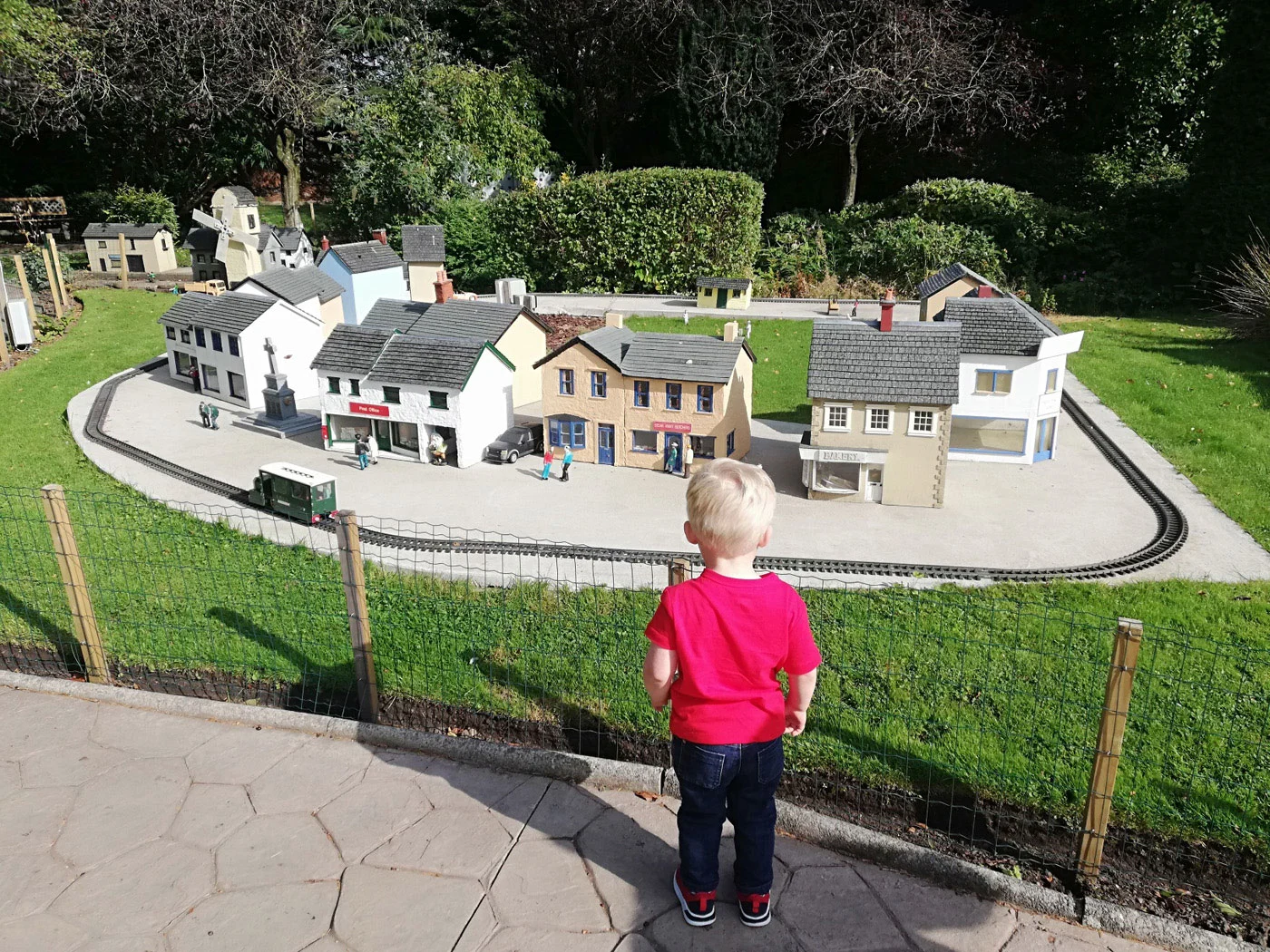 A young boy in a red t-shirt gazes at an outdoor model village with miniature buildings and a small railway track.