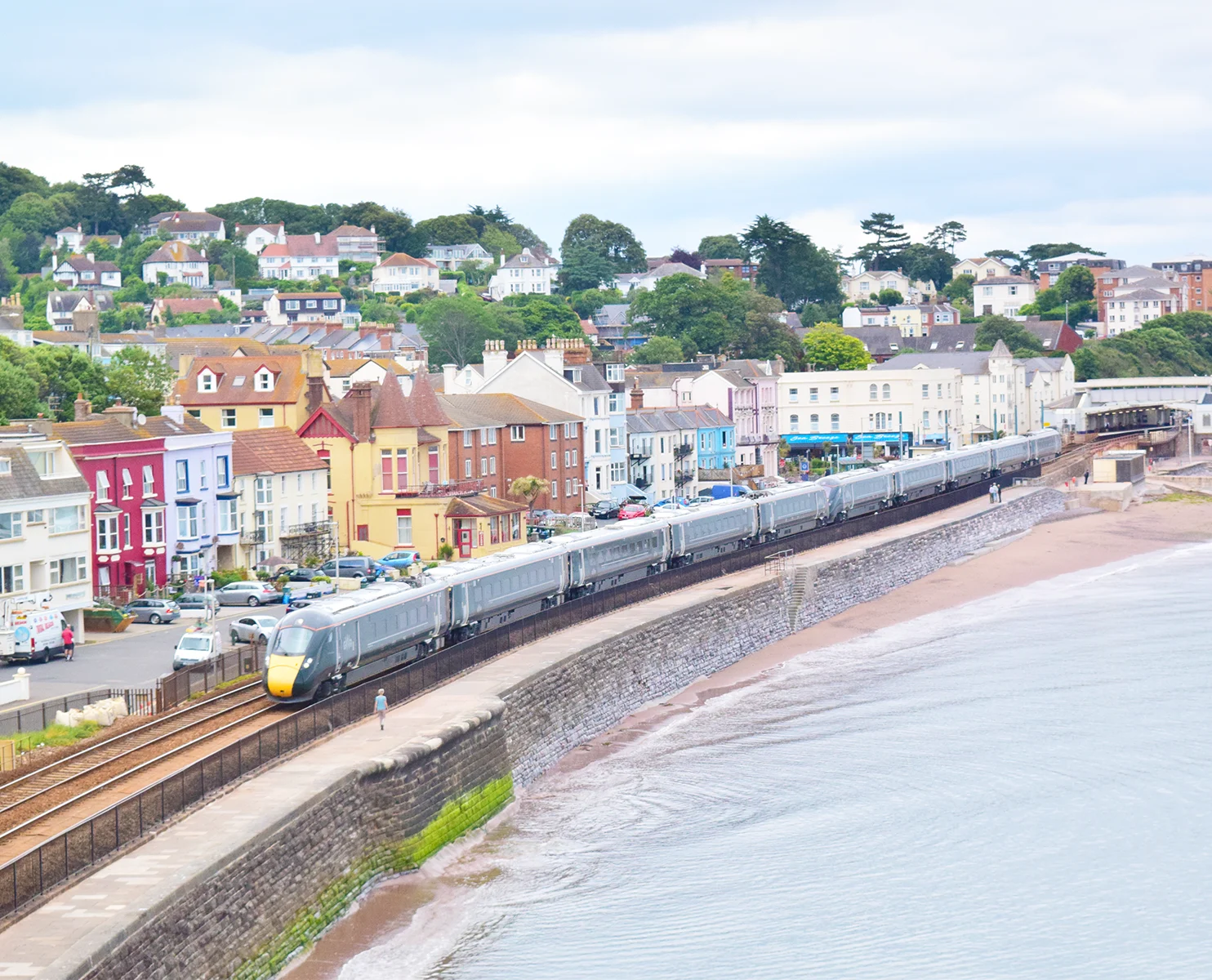 A train going along a track next to a beach with colourful houses and trees on the other side of the tracks.