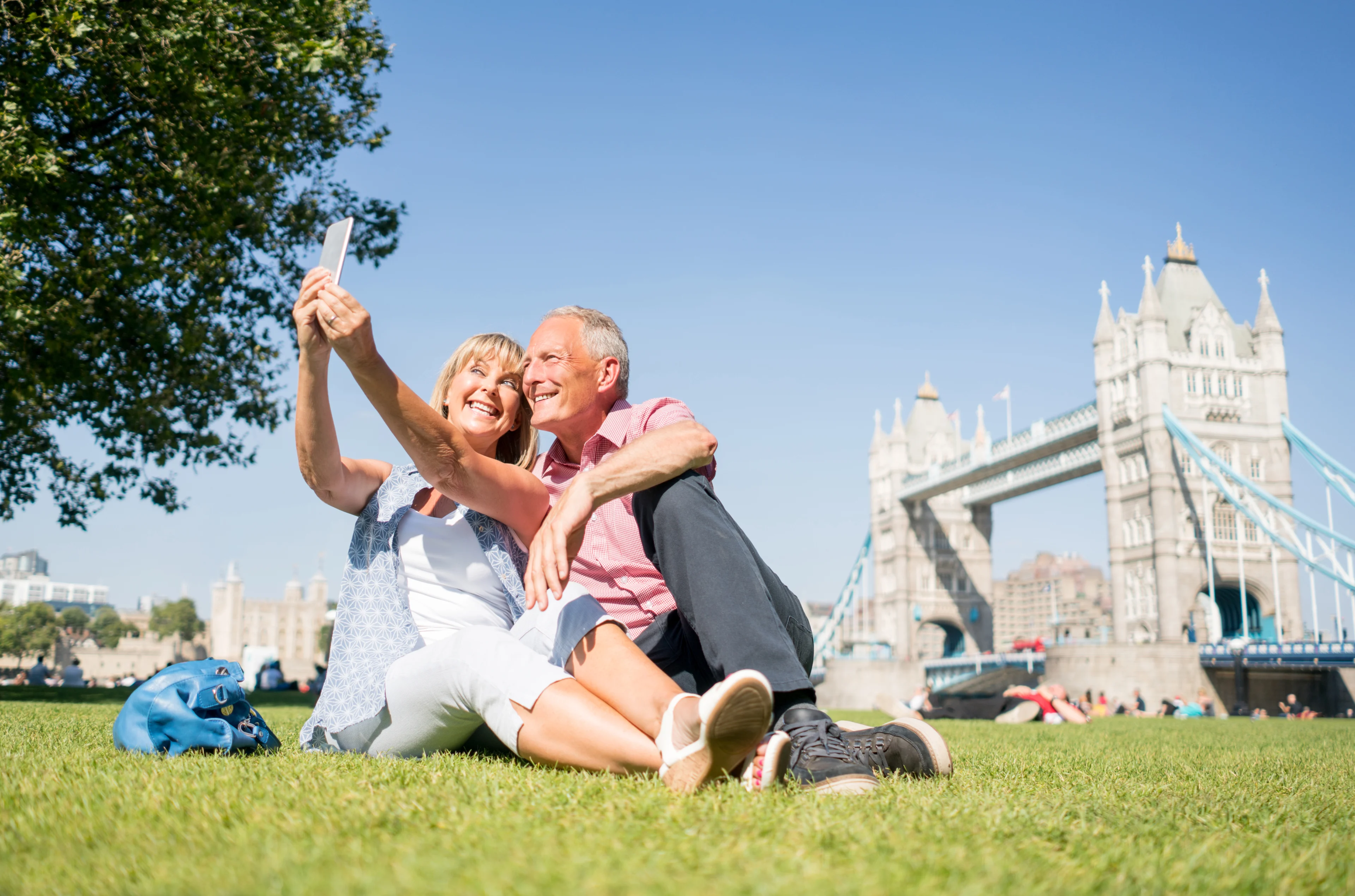 An older white man and woman relax on the grass and take a selfie with Tower Bridge in the background