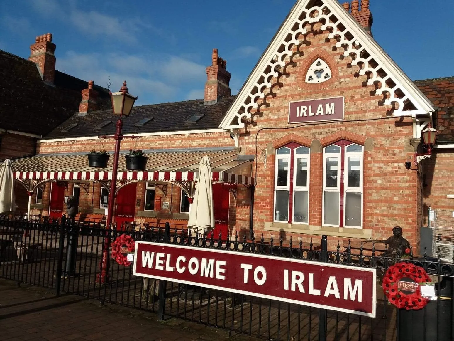A red brick Victorian railway station with a "Welcome to Irlam" sign, red and white striped awnings, and poppy wreaths on the fence.
