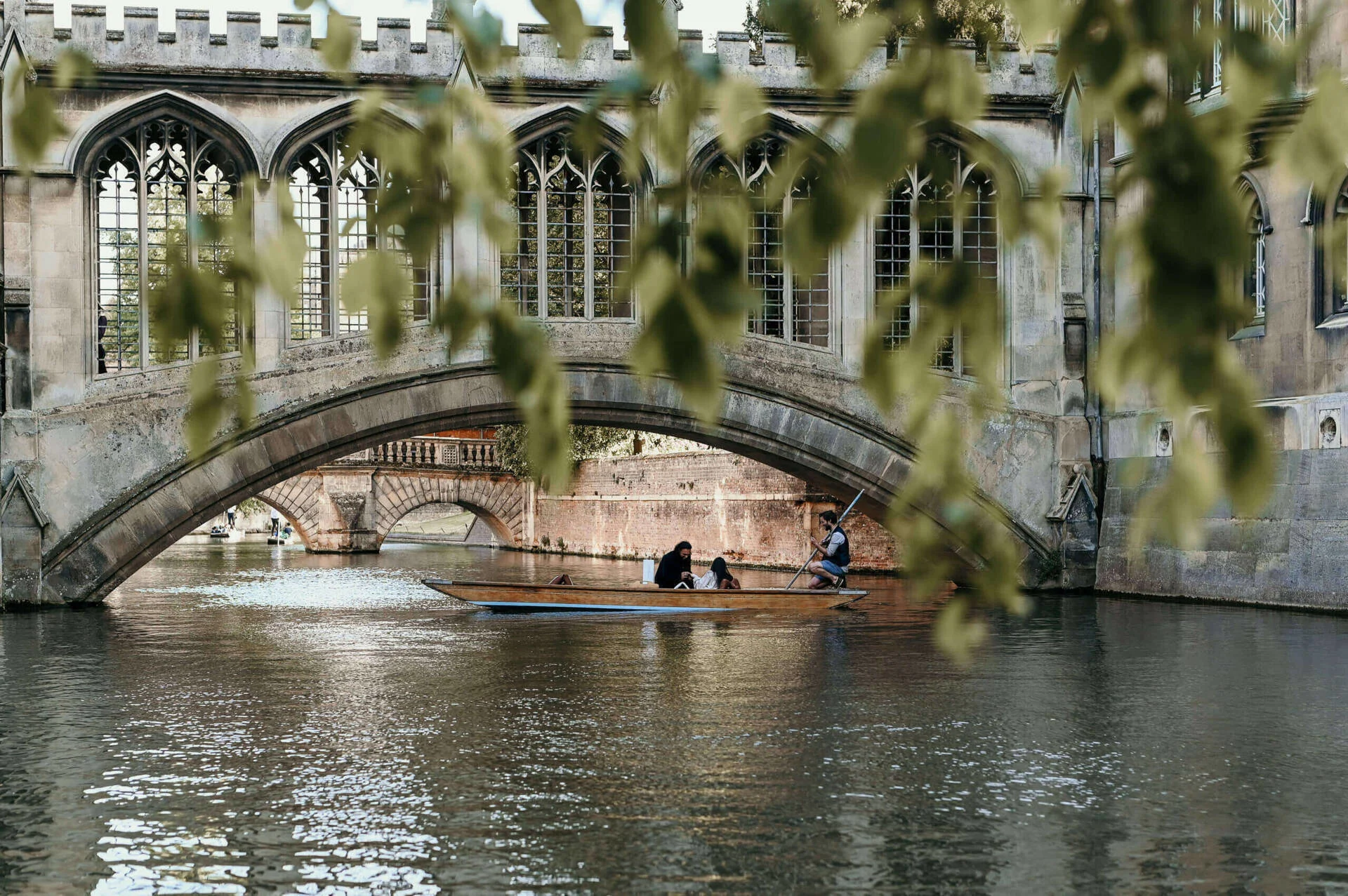 A couple sitting in a punt on a river, with a man holding a pole to push them along, they are going underneath a stone arch of an enclosed bridge.