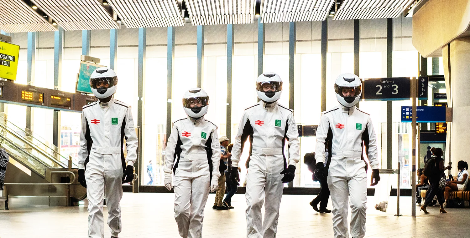 4 people dressed in white motor racing jumpsuits and wearing white racing helmets, walking side by side towards the camera  in the hall of a large railway station.