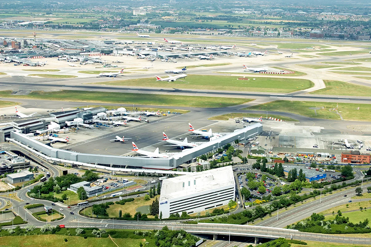 Aerial view of a large airport with planes waiting on the runways and green fields and trees in the distance. 