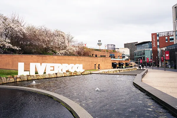 A river running by a white sign that says LIVERPOOL, with a paved street and a large shopping centre next to it. 