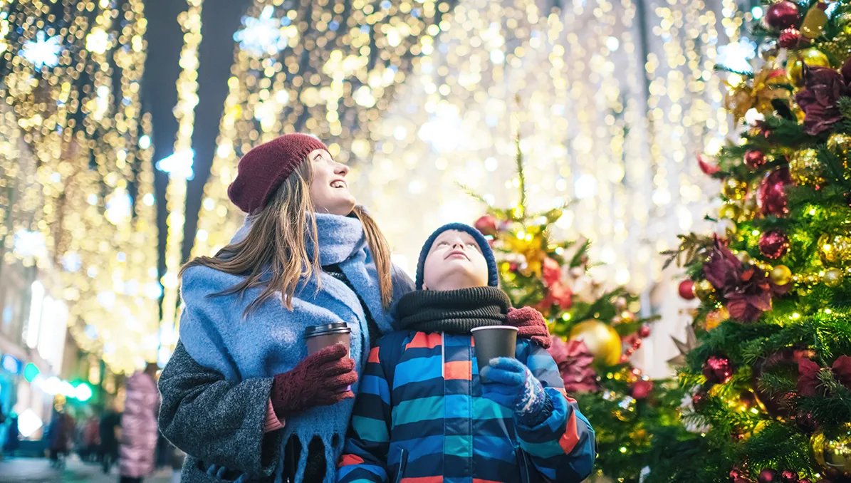 A white woman a white boy in winter coats, wooly hats and scarves, holding hot drinks and standing in front of a twinkling Christmas tree and huge blankets of lights. 