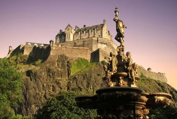Edinburgh castle sits on a hill top beneath a purple sunsetting sky. A statue of people in dark materials sits in the foreground.