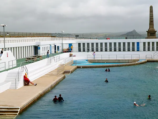 An outdoor lido with white walls, swimmers in the pool, a lifeguard seated nearby, and a stone obelisk visible in the background.