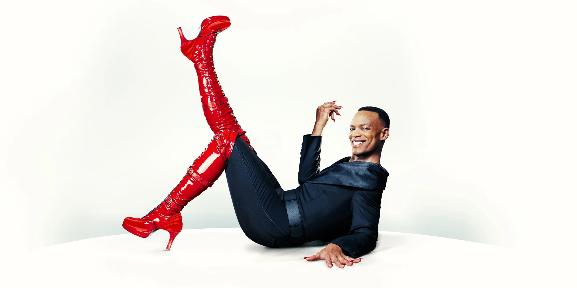 A Black person in a black outfit lying back and posing with one leg raised, wearing striking red thigh-high boots with high heels against a white background.
