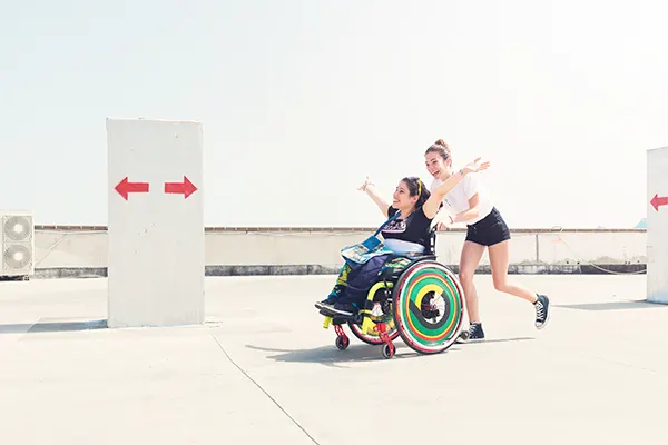 2 white women, 1 in a wheelchair and 1 pushing the wheelchair, smiling and laughing as they make their way along a rooftop car park.