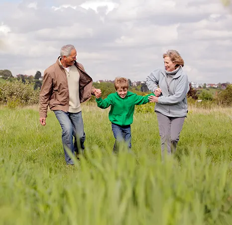 A senior white man and woman on either side of a young white boy, they are holding hands and smiling as they run through a green field. 