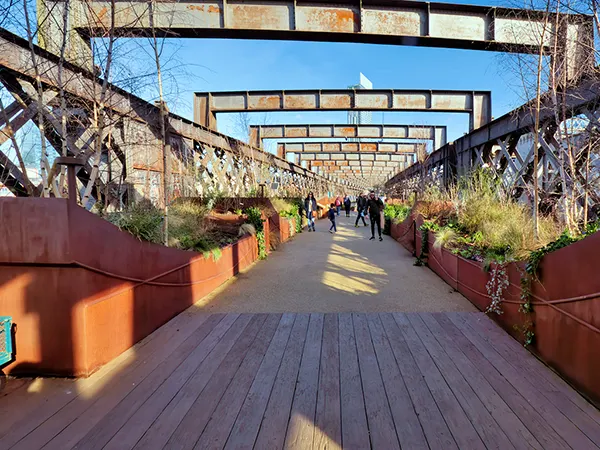 A wooden path on a railway viaduct with foliage in planters either side and large metal girders bridged across the top. The sky is blue and people are walking along in the background. 