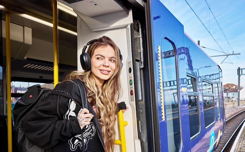 A young white woman with long blonde hair, wearing headphones and a backpack, stepping off a blue train at a railway station platform on a sunny day.