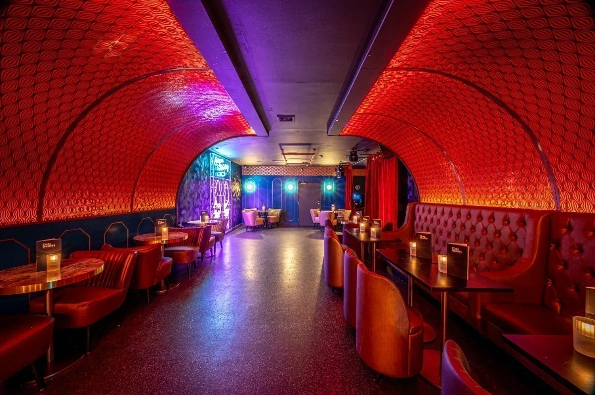 A dimly lit cocktail bar with red patterned arched walls, velvet seating, and purple and blue neon lighting.