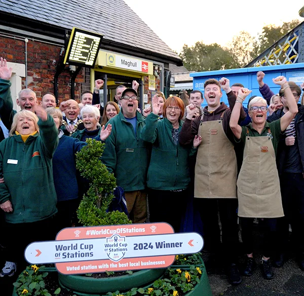 A group of station and cafe staff member wearing green fleeces and beige aprons, cheering and waving their arms. They are standing by a sign that says 'World Cup of Stations 2024 Winner' and in front of the station building with a sign that says 'Maghull'.