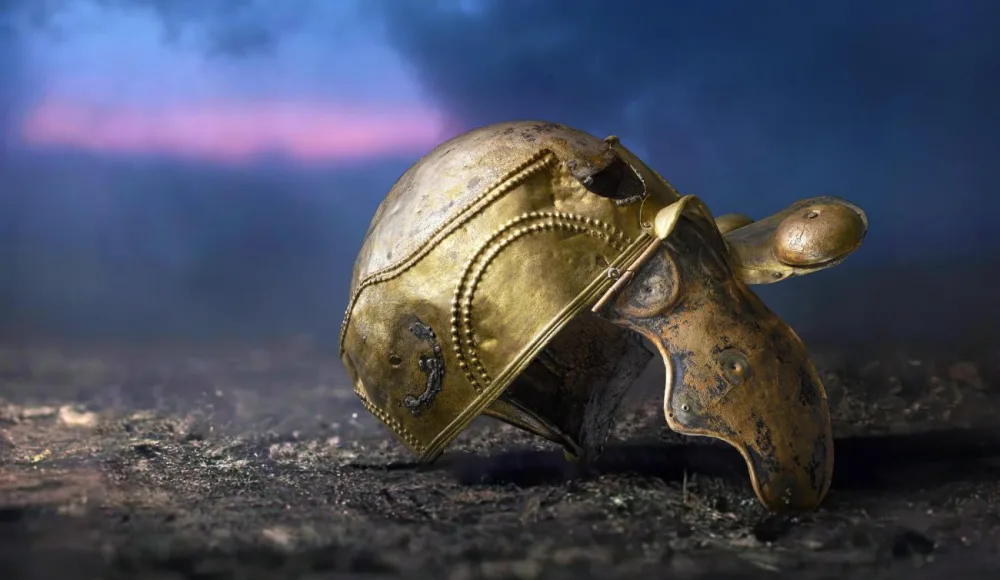 A gold ancient Roman helmet lying on soil, with a purple sky in the background. 