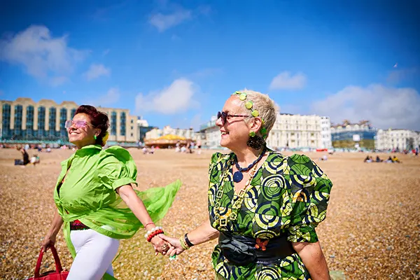 Two women in vibrant green outfits hold hands on a sunny pebble beach, smiling, with seaside buildings in the background.