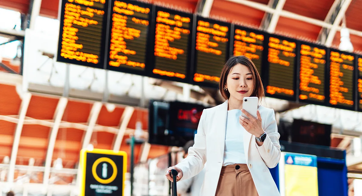 An East Asian woman standing in front of a large departure board at a rail station, looking at her phone and holding a wheeled suitcase.