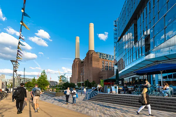 Riverside view of Battersea Power Station, a big red brick building with huge white chimneys, and glass fronted towers next door with shops at ground level. People are walking past on a sunny day.