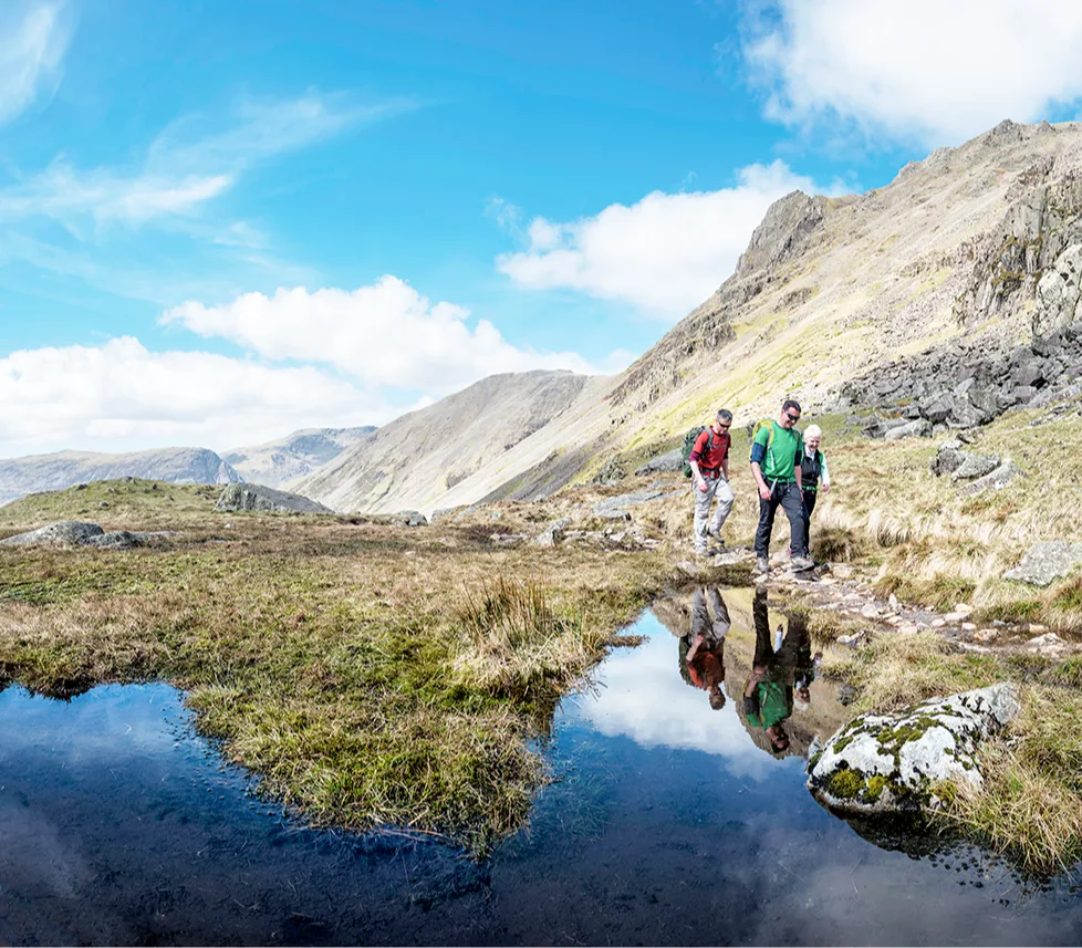 2 white men with brown hair and an older white woman with white hair, dressed in summer hiking gear and walking next to a lake in rugged, hilly countryside. 