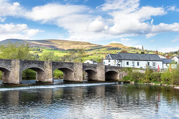 A stone bridge over a river with rolling hills in the background and a white building with a grey slate roof on the opposite side of the river.
