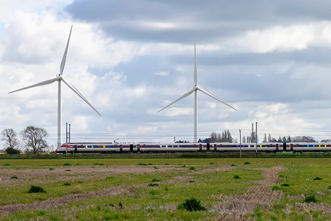 A train travelling across a green field during the day, with large white wind turbines in the background. 